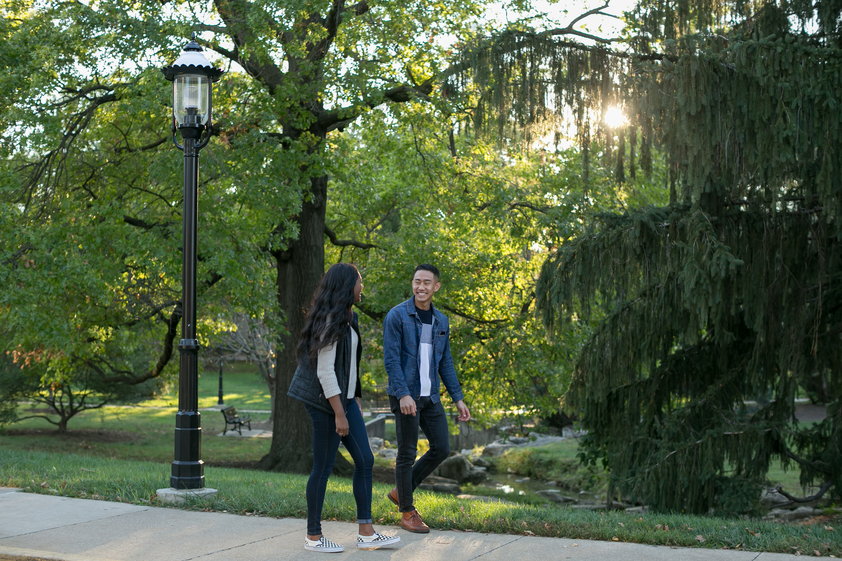 A young man and woman walk along a tree-lined path, smiling and talking. The sun filters through the trees, creating a warm, relaxed atmosphere.